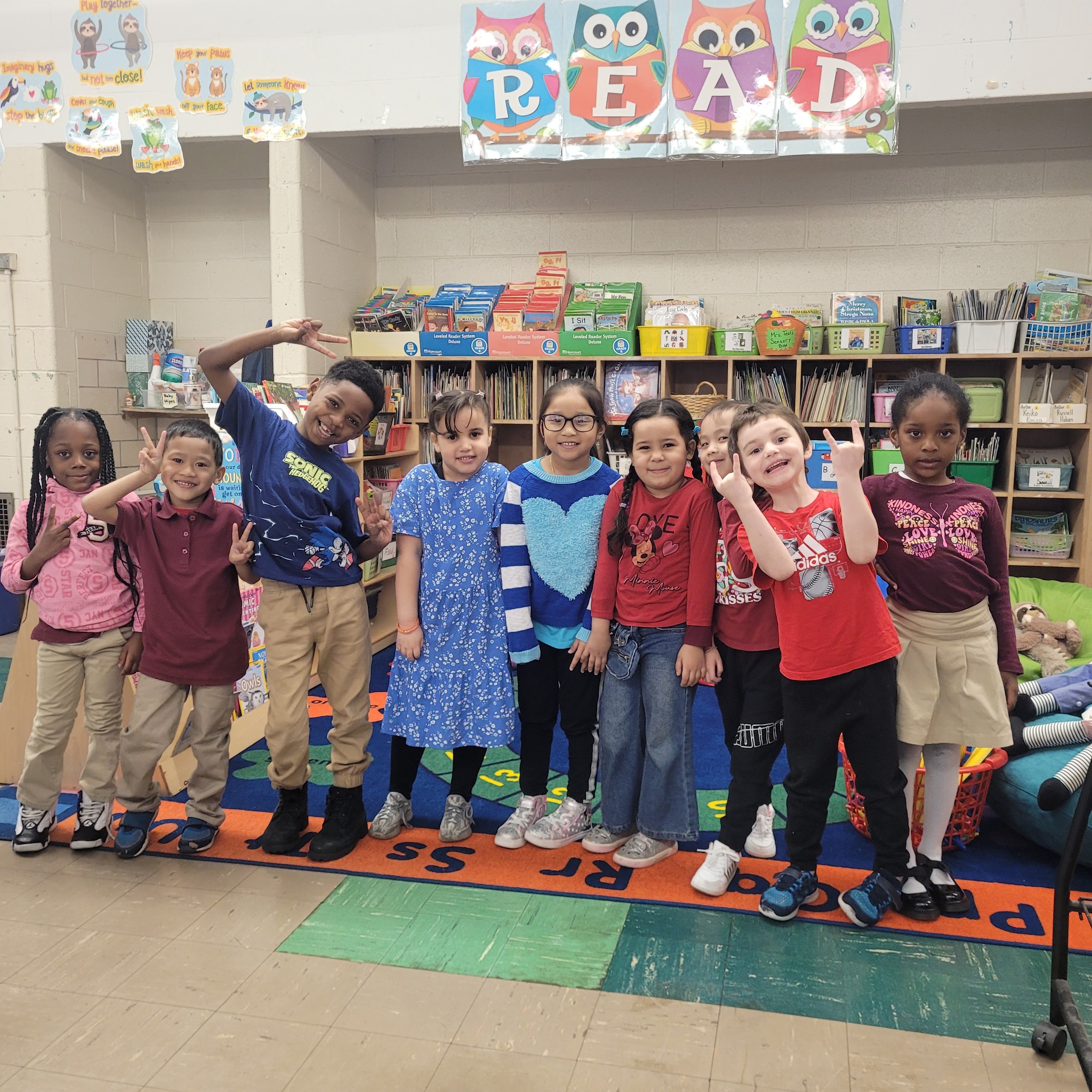 A group of smiling students stand together on a colorful classroom rug at Stephen Girard Elementary School. Behind them, a bookshelf and a “READ” banner decorate the space, creating a warm and welcoming reading corner.
