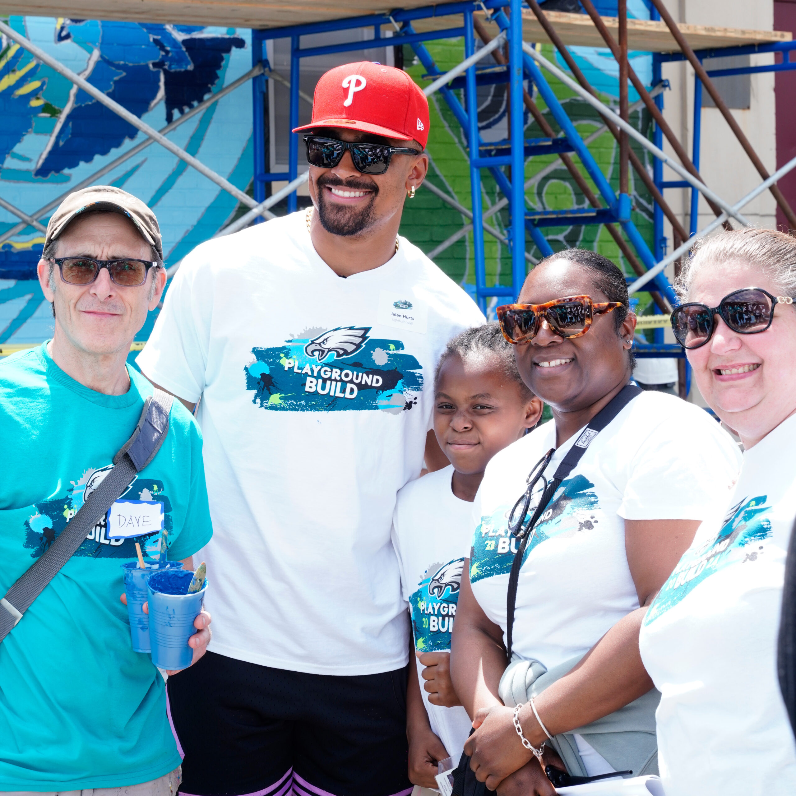 A group of volunteers smile together during the Philadelphia Eagles Playground Build at Stephen Girard Elementary School in Philadelphia. They’re standing in front of a colorful mural, wearing event T-shirts and sunglasses, celebrating a successful day of community service.