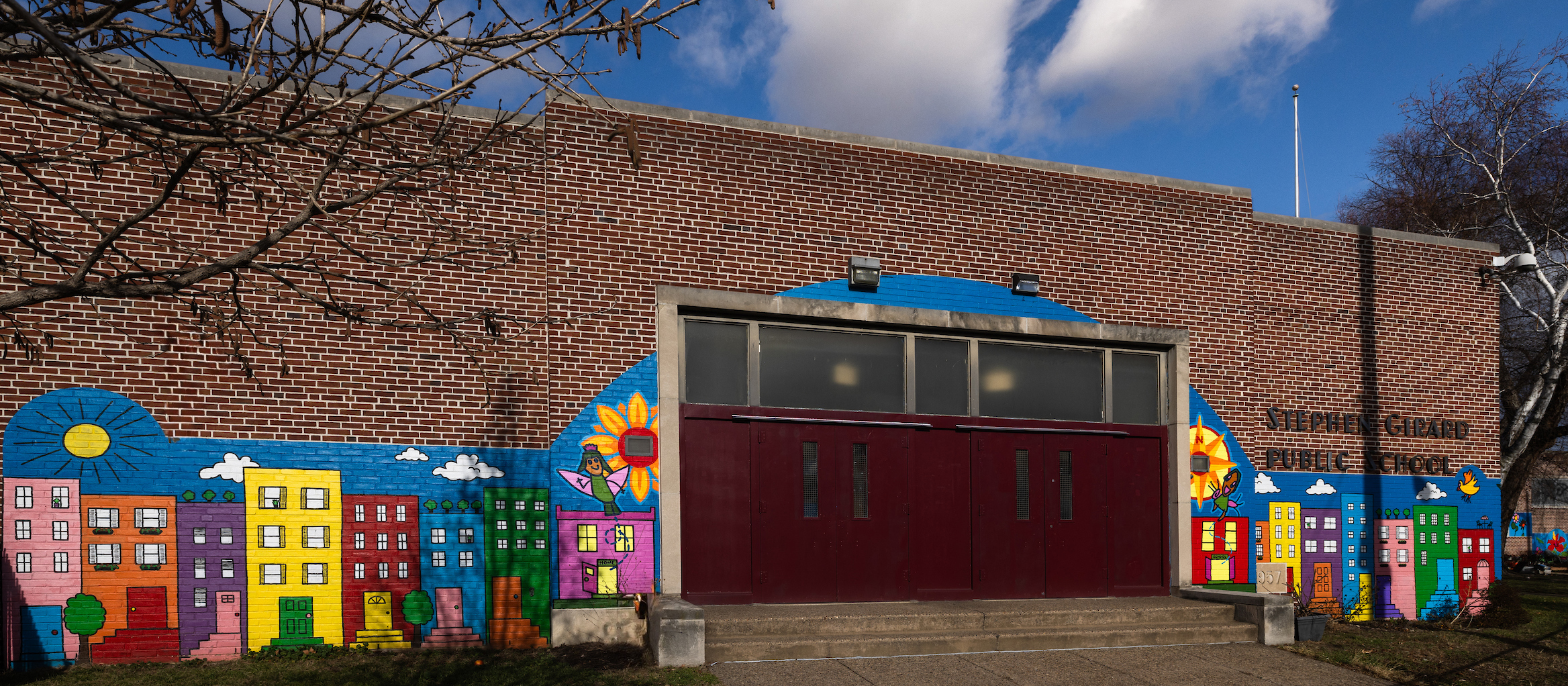 A vibrant mural on the front of Stephen Girard Elementary School shows a row of colorful city buildings, a sun, and playful illustrations painted on the brick wall,.
